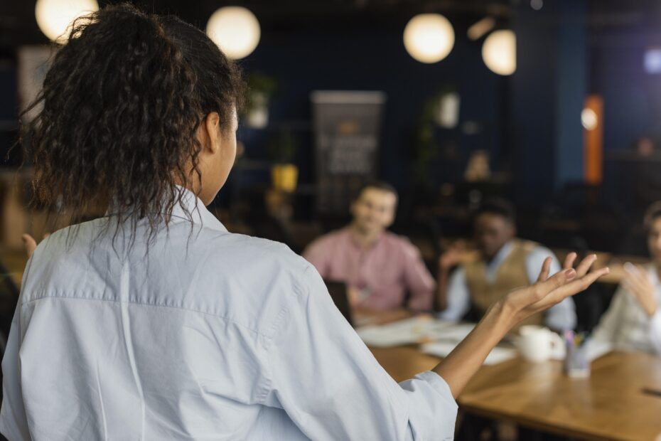 Imagem de Freepik mostra mulher de costas para a foto gesticulando durante sua fala para pessoas que estão sentadas numa mesa