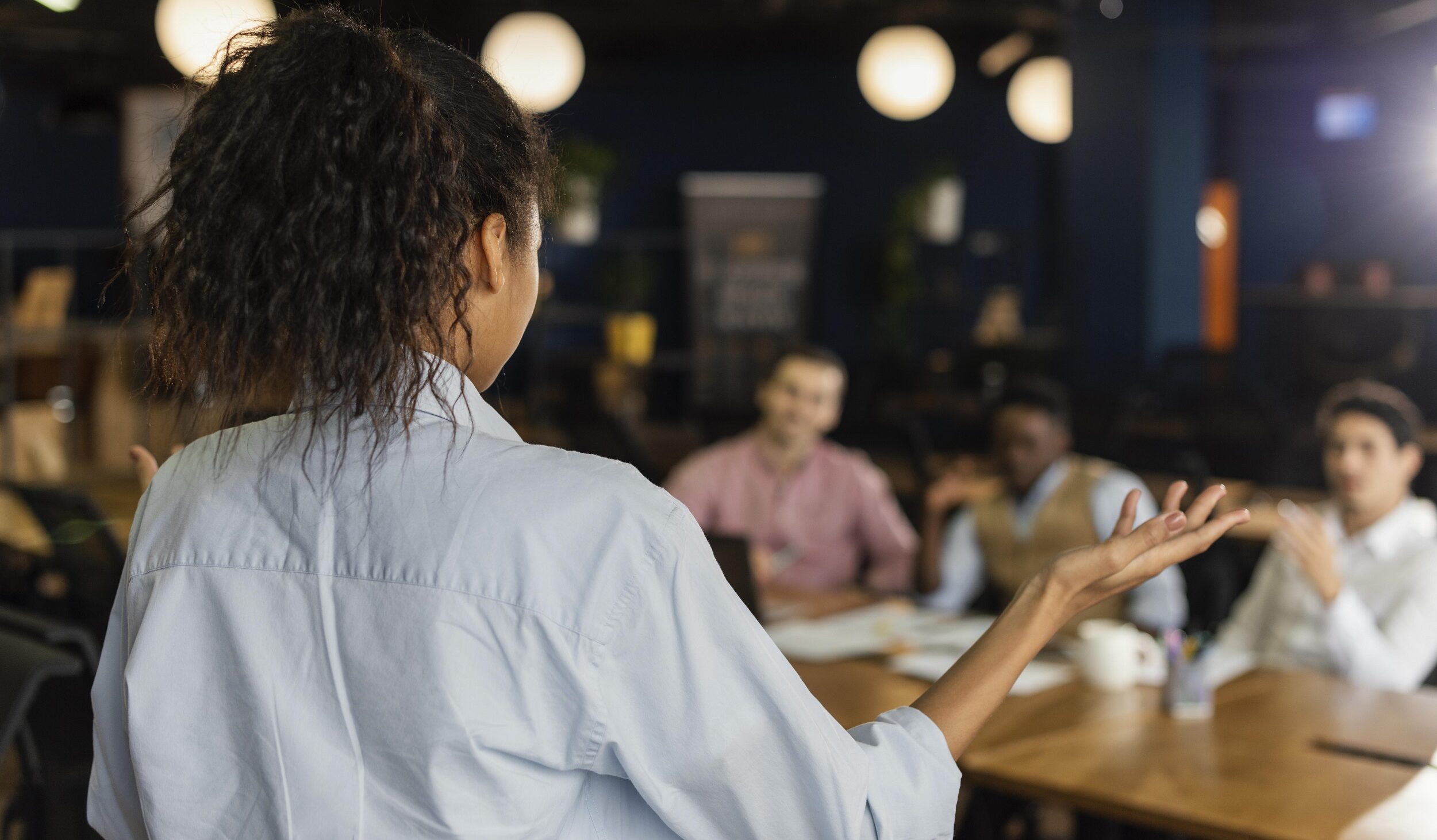 Imagem de Freepik mostra mulher de costas para a foto gesticulando durante sua fala para pessoas que estão sentadas numa mesa