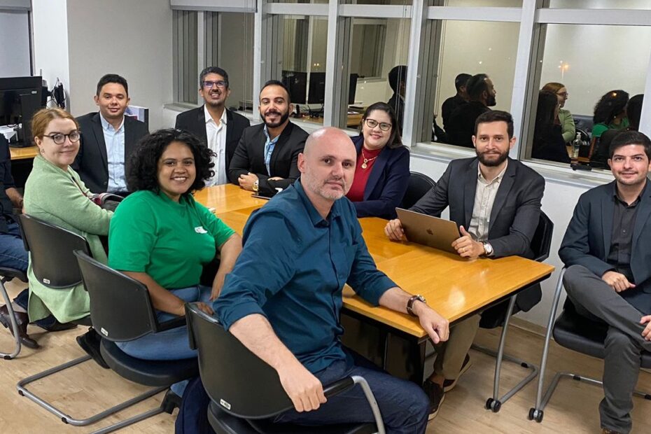 Representantes do NEES sentados ao redor de uma mesa retangular amarela, todos olham para a foto