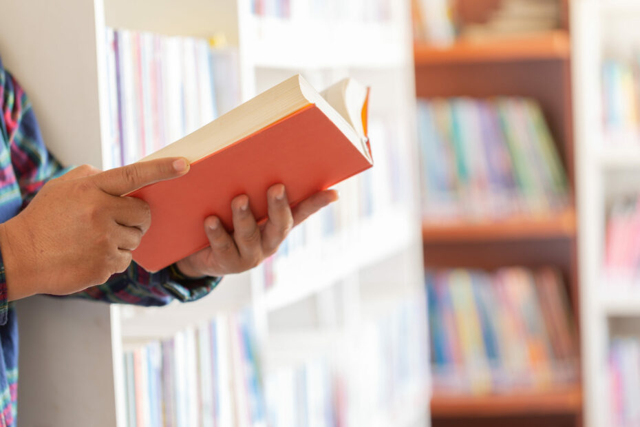 Pessoa lendo um livro de capa laranja. Veste camisas de mangas longas. O fundo desfocado mostra prateleiras de livros