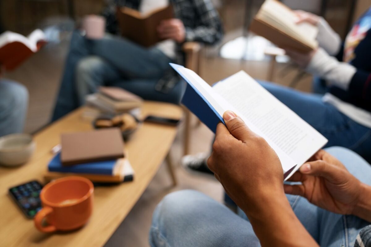 Pessoa segurando livro pequeno de capa azul. Mãos, livro e uma mesa pequena aparecem