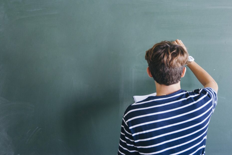 Estudante vestindo camisa azul com listras brancas horizontais, está em pé escrevendo com giz num quadro negro e segurando um caderno