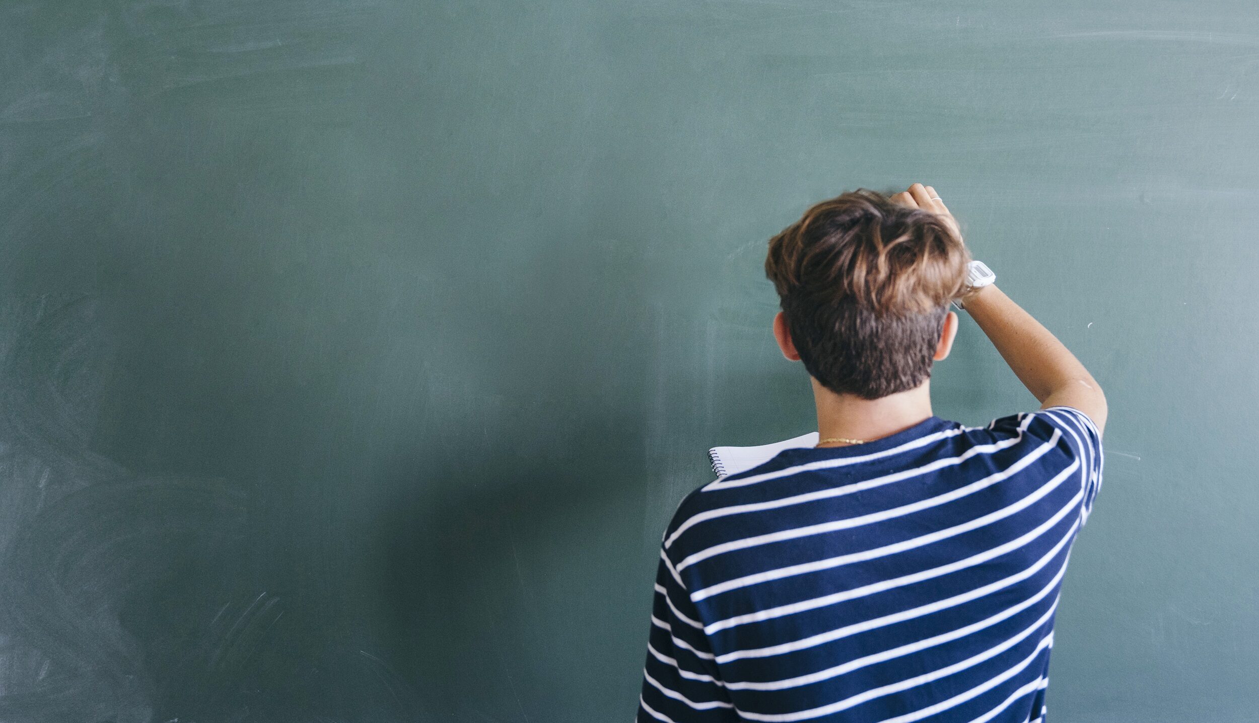 Estudante vestindo camisa azul com listras brancas horizontais, está em pé escrevendo com giz num quadro negro e segurando um caderno